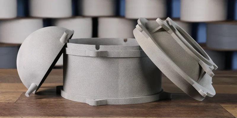 Three grey 3D-printed PA12 nylon parts on a workbench — a cylindrical housing with a domed cover and a ribbed circular lid, against a blurred rack of filament spools